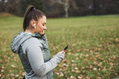 Akıllı telefonunu kullanan ve dışarıda parkta egzersiz yaparken kablosuz kulaklıkla müzik dinleyen odaklanmış genç bir kadın..