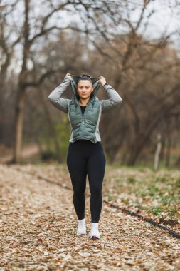 Yeşil ceketli bir kadın kafasına bir kapüşon geçirir ve sonbaharda parkta yaprak kaplı bir patikada koşuya hazırlanır..
