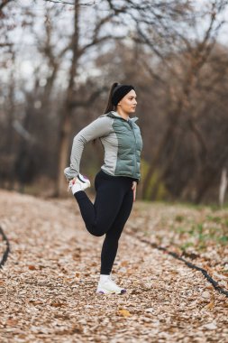 Odaklanmış genç bir bayan koşucu, şehir parkında yaprak kaplı bir patikada ayakta durarak koşuyor..
