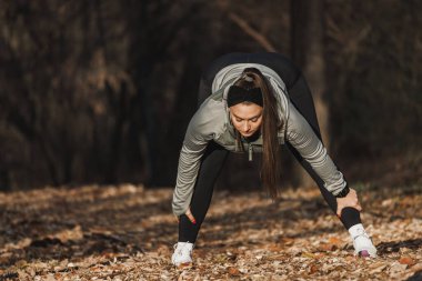 Aktif kıyafet giyen odaklanmış genç bir kadın antrenmana hazırlanmak için parkta esneme hareketleri yapıyor..