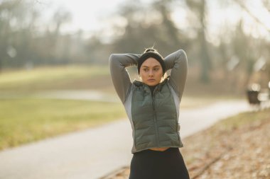 Aktif kıyafet giyen odaklanmış genç bir kadın antrenmana hazırlanmak için parkta esneme hareketleri yapıyor..