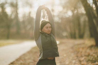 Odaklanmış genç bir kadın, güneş lekeli bir yolda kollarını uzatarak kendini enerjik bir sabah koşusuna hazırlıyor..
