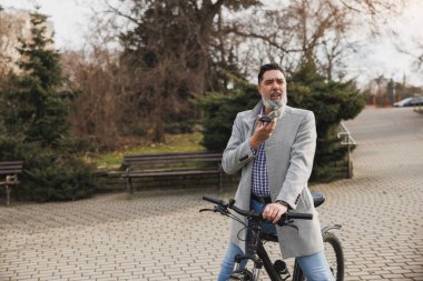 Sakallı sıradan bir iş adamı şehir parkındaki bir bisikletin tepesinde oturmuş cep telefonuna sesli mesaj kaydediyor..