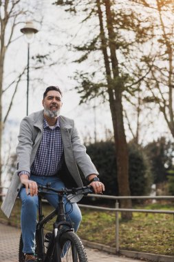 Sakallı sıradan bir iş adamı şehir parkında bisiklet sürüyor ve önüne bakıyor..