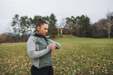 Odaklanmış genç bir kadın akıllı hareketlerini kontrol etmek için durur ve parkta egzersiz yaparken ilerlemesini izler..