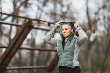 Yeşil ceketli bir kadın kafasına bir kapüşon geçirir ve kışın parkta koşuya hazırlanır..