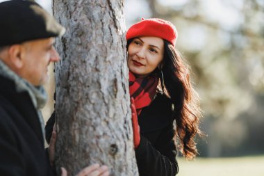 Kırmızı bereli orta yaşlı bir kadın bir ağacın arkasından, sonbahar boyunca parktaki olgun kocasına bakıyor..