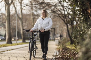 Genç, gülümseyen bir kadın bisikletini kaldırımda yürüyor ve etrafa bakıyor..