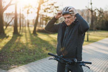 Kasklı yaşlı bir adam şehir parkında bisiklet sürmeye hazır..