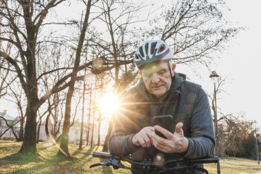 Son sınıf öğrencisi gün batımında şehir parkında yanında motosikletle cep telefonuna odaklanmış duruyor..
