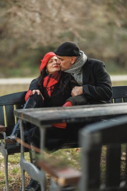 Sevecen, orta yaşlı bir çift sarılıp öpüşürken güneşli bir kış gününde bir bankta rahatça oturuyorlar..