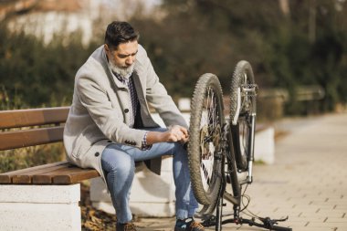 Sakallı biri bisiklet lastiğini tamir etmeye odaklanır. Parktaki ahşap bir bankta oturuyordu. Bisikleti ters dönmüştü..