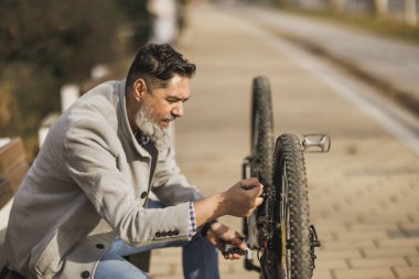 Sakallı bir adam devrilmiş bisikletini tamir ederken bir bankta oturuyor, kentsel çevrede tamir aletleri kullanıyor..