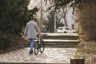 Sıradan bir iş adamının arka görüntüsü, dağ bisikletiyle şehir parkında bir dizi taş basamağa tırmanıyor..