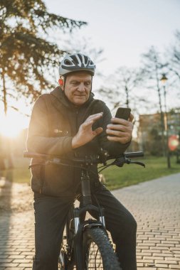 Kask takan yaşlı bir adam, güneşli bir parkta bisikletine yaslanırken akıllı bir telefonla video görüşmesi yapıyor..