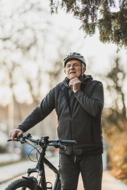 Son sınıf öğrencisi bir adam şehir caddesinde bir bisikletin yanında duruyor. Bisiklet kaskını ayarlar ve güvenli bir bisiklet sürüşe hazırlanır..