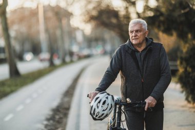 Yaşlı bir adam kaldırımda bisikletinin yanında yürüyor ve etrafa bakıyor..