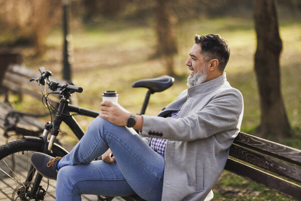 A relaxed middle-aged man is seated on a park bench, taking a moment to coffee rest while outdoors. His bike is parked beside him.