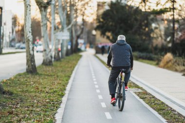 Yaşlı bir adamın arka manzarası güneşli bir günde ağaçlarla çevrili bir şehir caddesinde bisiklet sürmenin keyfini çıkarıyor..