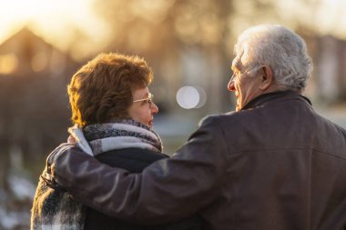 Mutlu, yaşlı, kucaklaşan bir çiftin şehirde yürüyüşe çıkışının arka görüntüsü..
