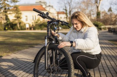 Genç bir kadın şehir parkının kaldırımında diz çökerken siyah dağ bisikletini yakından inceliyor..