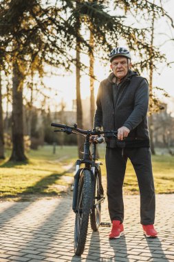 Kasklı son sınıf öğrencisi, güneşli bir şehir parkında kaldırımda dağ bisikletinin yanında duruyor. Kameraya bakıyorum..