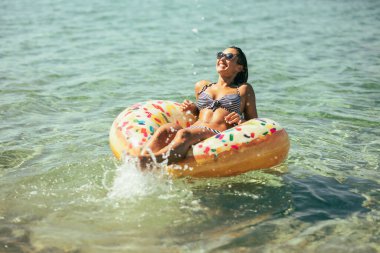 Bir kadın şişme halka bir çöreğe biniyor ve yaz tatilinin tadını çıkarırken su sıçratıyor..