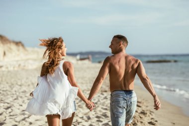 Bir çiftin arka görüntüsü deniz kıyısındaki kumlu sahilde yan yana koşuyor. Rahatlamış görünüyorlar ve güneşli bir günün tadını çıkarıyorlar..