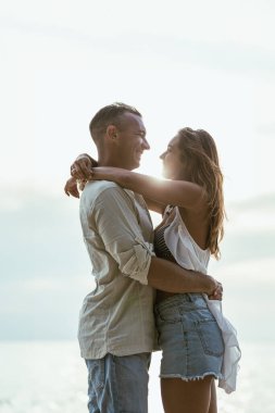 Bir adam ve bir kadın arka planda okyanus olan kumlu bir plajda birbirlerine sarılıyorlar..