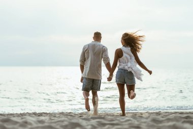 Arka planda mavi deniz ve kumlu sahil boyunca el ele koşan bir çiftin keyfini çıkarıyorlar..