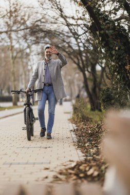 Sakallı sıradan bir iş adamı bisikletinin yanında rahatça yürüyüp dostça bir jest yaparak elini sallıyor..