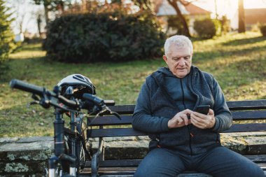 Üst sınıftan bir adam parktaki bankta otururken sosyal medyada akıllı telefonuyla sörf yapıyor. Bisikleti onun yanına park edilmiş..