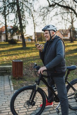 Kask takan son sınıf öğrencisi, şehir parkında bisiklet sürerken cep telefonundan sesli mesaj gönderiyor..