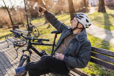 Kask takan orta yaşlı bir adam, parkta bir bankta mola verirken akıllı telefonda selfie çekiyor. Bisikleti onun yanına park edilmiş..