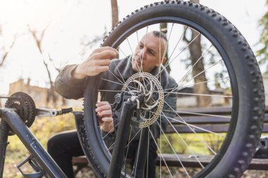 Orta yaşlı bir adam tahtadan bir bankta otururken devrilmiş bisikletini tamir etmeye odaklanır..