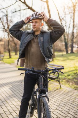 Orta yaşlı bir bisikletçi şehir parkında dağ bisikletiyle dururken kask takıyor..