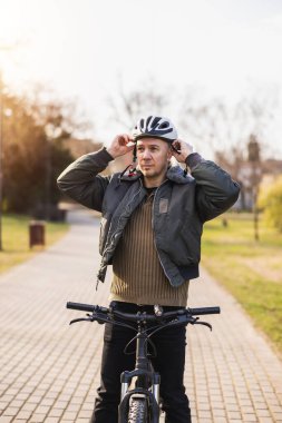 Miğfer takan orta yaşlı bir bisikletçi şehir parkında dağ bisikletiyle duruyor, güvenli bir yolculuğa çıkmaya hazır ya da bir tanesini yeni bitirmiş..