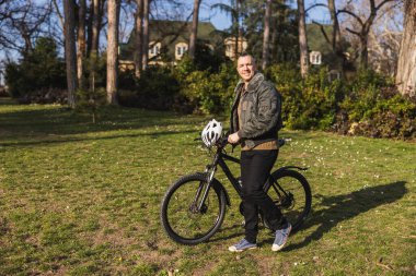 Orta yaşlı bir adam bisikletiyle yürüyor ve güneşli bir sonbahar gününde şehir parkında günün tadını çıkarıyor..