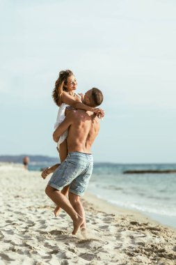 Kumlu sahil boyunca yürürken eğlenen bir çift. Adam kadını tutuyor, kolları boynuna dolanıyor..