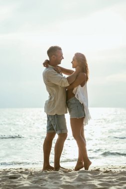 Arka planda mavi deniz olan kumlu bir plajda bir çift birbirini kucaklıyor..