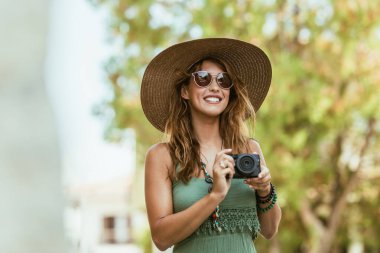 Şapka takan ve güneş gözlüğü takan gülümseyen bir kadın elinde bir kamera tutuyor, yaz tatilinde anı kaydetmeye hazır..