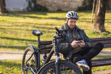 Kask takan orta yaşlı bir adam park halindeki bir bankta oturuyor, park halindeki bir bisikletin yanında, cep telefonuyla sosyal medyada geziniyor..
