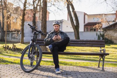 Gülümseyen orta yaşlı bir adam, parktaki bankta oturuyor ve kendini cep telefonuna kaptırmış, daktilo ediyor ve kayıyor. Bisikleti onun yanına park edilmiş..