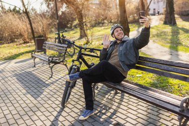 Gülümseyen orta yaşlı bir adam parktaki bir bankta park halindeki bir bisikletin yanında otururken akıllı telefonuyla bir video görüşmesi yapıyor..