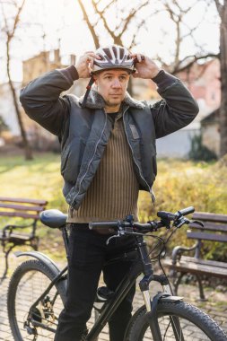 Orta yaşlı bir bisikletçi, kasklı bir bisikletin yanında dikilir. Güvenli bir sürüş için hazırdır ya da yeni bitirmiştir..