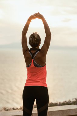 Güneşin doğuşunda kollarını başının üstüne uzatan kaslı bir kadının arka görüntüsü. Yaz tatilinde bir plaj kenarında yoga antrenmanı yapıyor..