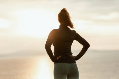 Güneşin doğuşunda rahatlayan kaslı bir kadının arka görüntüsü. Yaz tatilinde deniz kenarında koşuya hazırlanıyor..