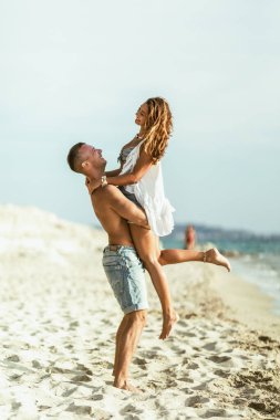Kumlu sahil boyunca yürürken eğlenen bir çift. Adam kadını tutuyor, kolları boynuna dolanıyor..