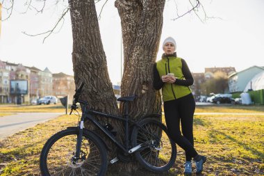 Bir kadın parkta bir ağaca yaslanmış duruyor, bisikletinin yanında. Akıllı telefonunu kullanıyor ve bisiklete ara veriyor..