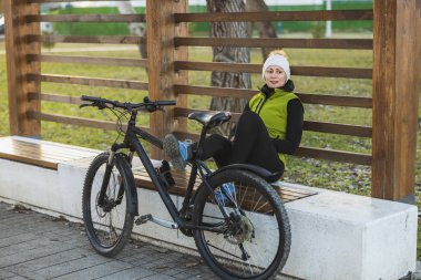 Rahat bir kadın park halindeki bir bisikletin yanındaki bankta oturuyor, çevreye bakıyor..